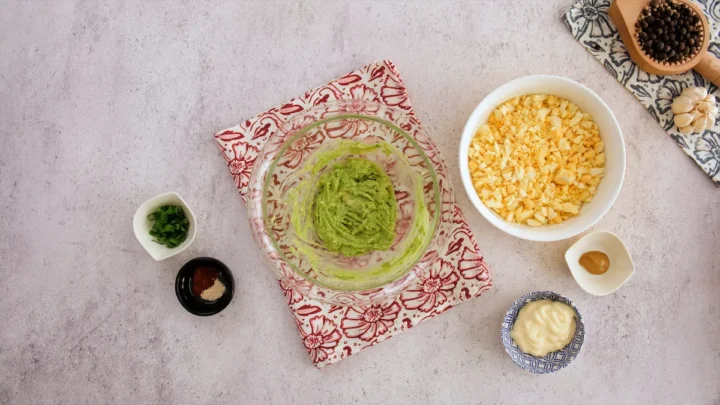 Mashed avocado beside a bowl of chopped hard-boiled eggs, mayo and other ingredients is on the table.
