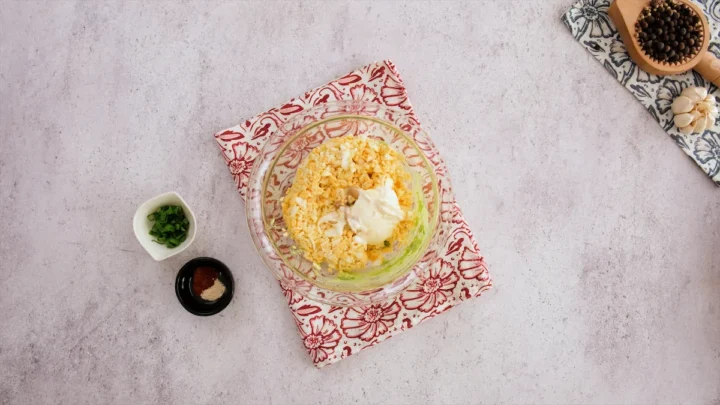 Eggs, mayo, and Dijon mustard added to mashed avocado in a mixing bowl, while spices and herbs are in small bowls on the table.