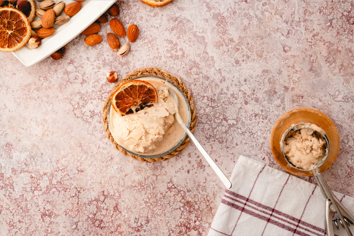 Homemade pecan butter ice cream topped with a slice of dried orange served in a glass bowl with a spoon alongside an ice cream scooper with more ice cream.