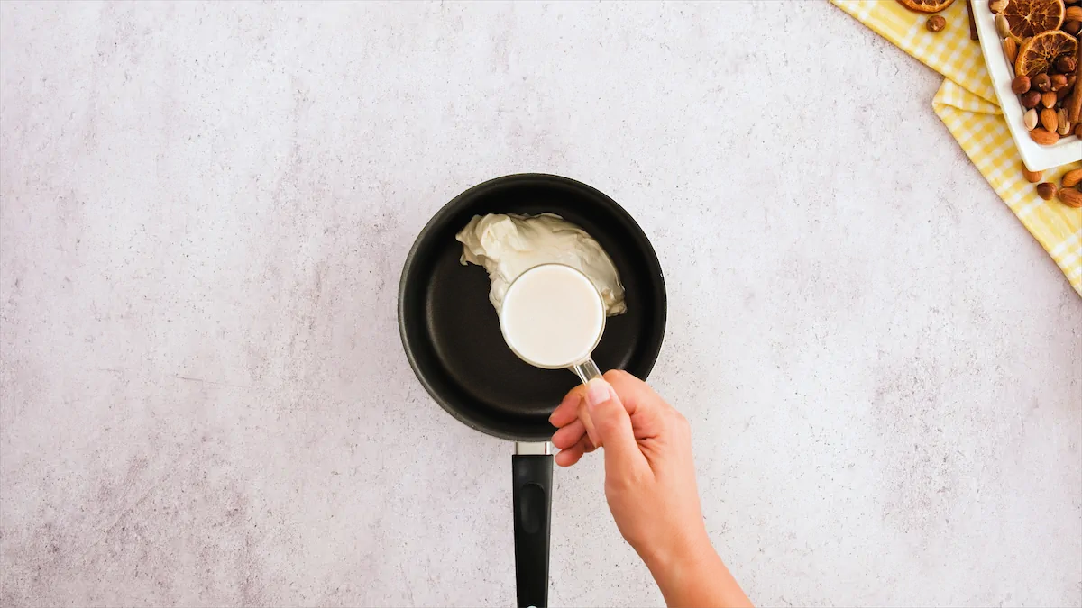 A hand holds a measuring cup filled with almond milk, which is ready to be added to the heavy cream in a saucepan.
