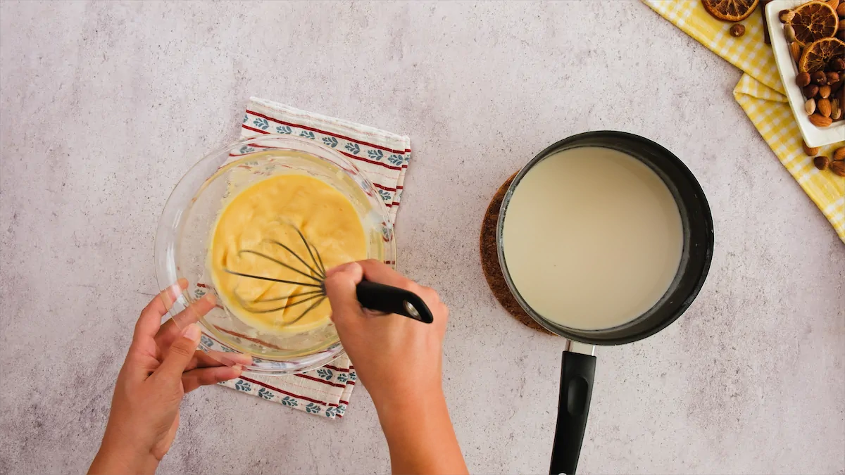 A hand is whisking together egg yolk and powdered erythritol, while beside it, a saucepan contains a heated mixture of heavy cream and almond milk.