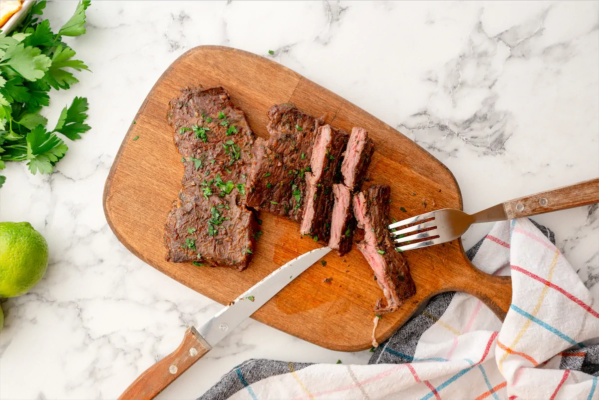 Two steaks of low-carb carne asada on a chopping board where one of the steaks is cut using a fork and knife.