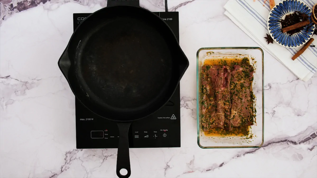 The cast iron skillet to cook carne asada is getting heated on an induction stovetop beside the ready-to-cook steaks in marinade.