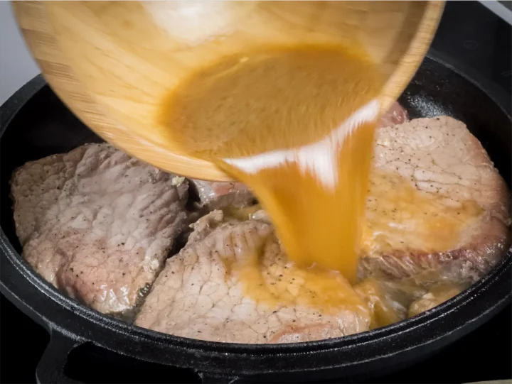 The sauce mixture is being added to the pork steaks cooking in a cast iron skillet.