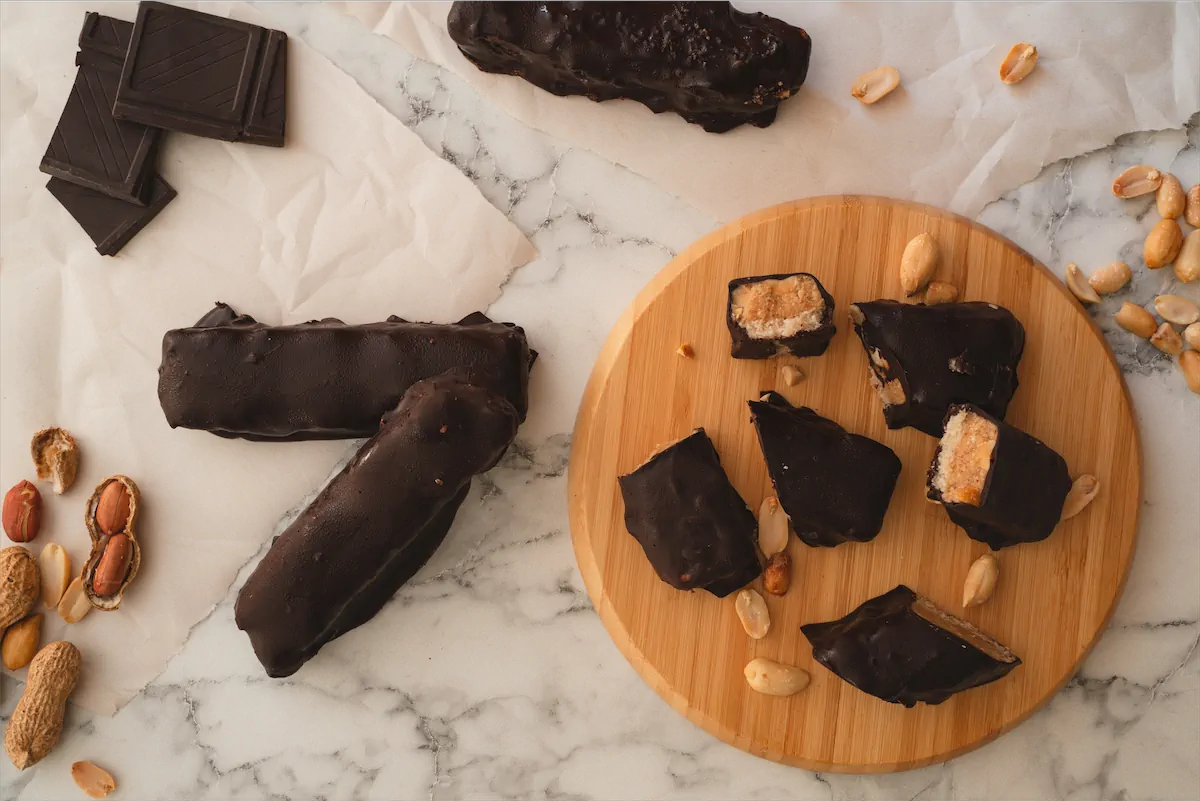 Homemade keto Snickers bars laid out on a table and some bars sliced to reveal their layers on the round wooden board.