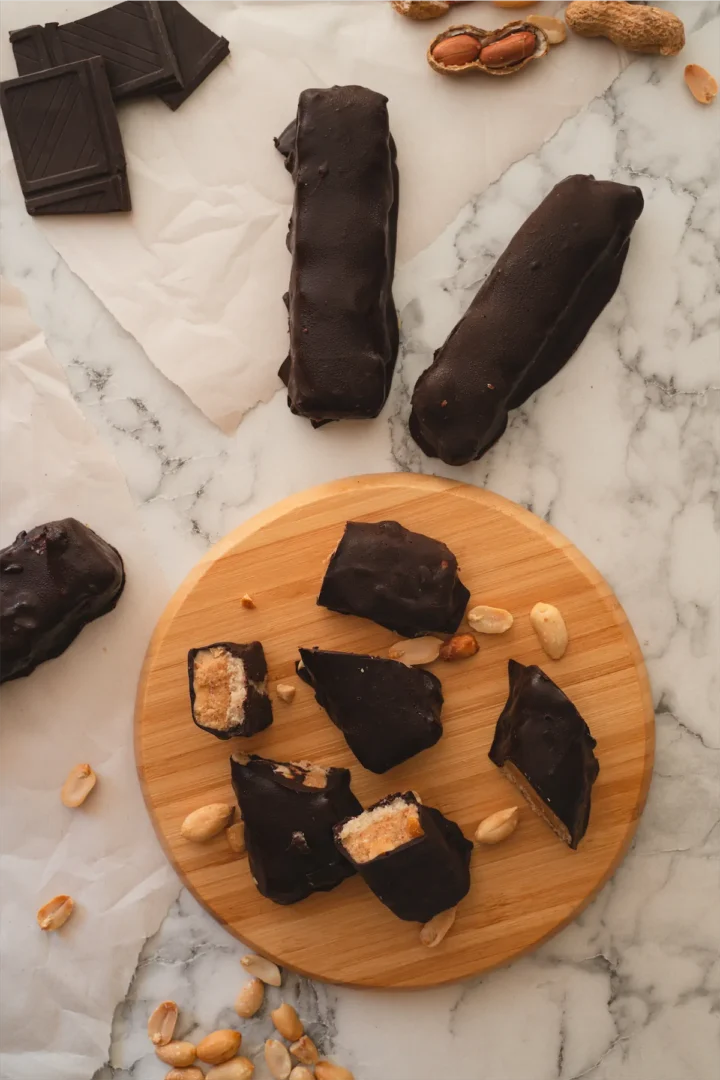 Keto copycat Snickers bars laid out on a table and some bars sliced to reveal their layers on the round wooden board.