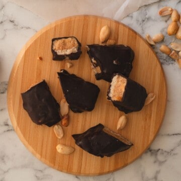 Keto Snickers bars cut showing its layers on a round wooden board and topped with peanuts.