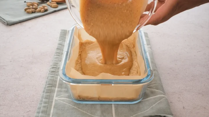 The caramel mixture getting poured over the frozen nougat layer in a freezer-safe glass container.