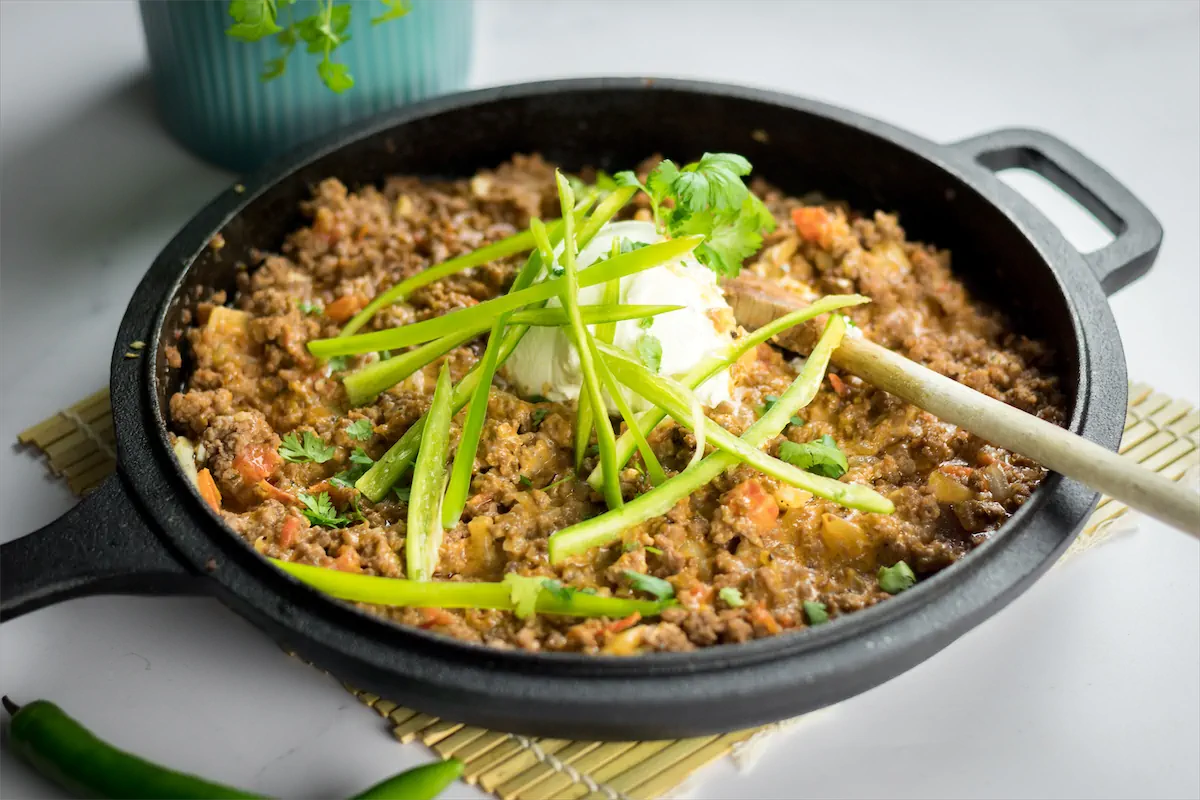 Homemade taco casserole is in a skillet, garnished with sour cream and fresh herbs along with a wooden spoon for serving.
