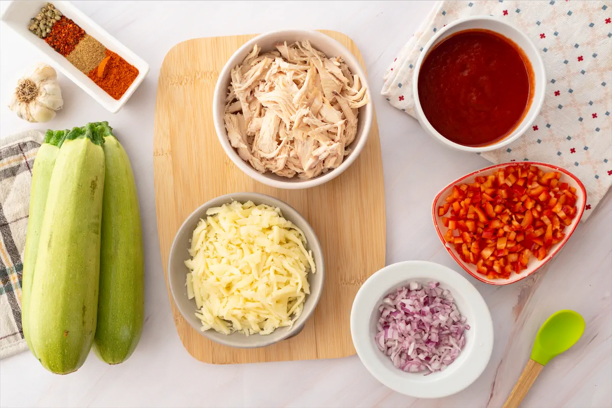 Zucchini, shredded cooked chicken, tomato purée, shredded mozzarella cheese, diced red onions and tomatoes, and spices gathered and arranged on the table.