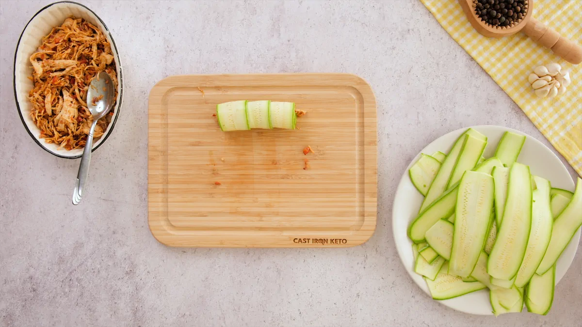 Zucchini strips, filled with shredded chicken, are placed seam-side down on a wooden board next to a plate of zucchini strips and a bowl of chicken filling.