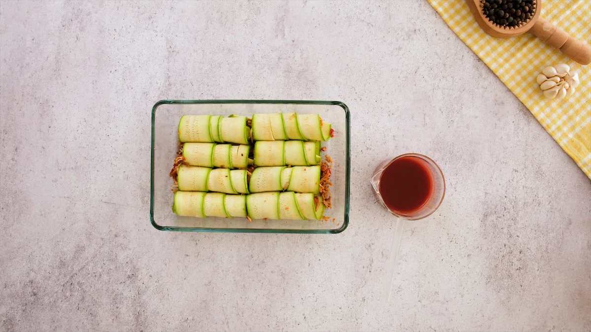 Rolled zucchini rolls are arranged in a baking dish beside the required amount of tomato sauce on the table.