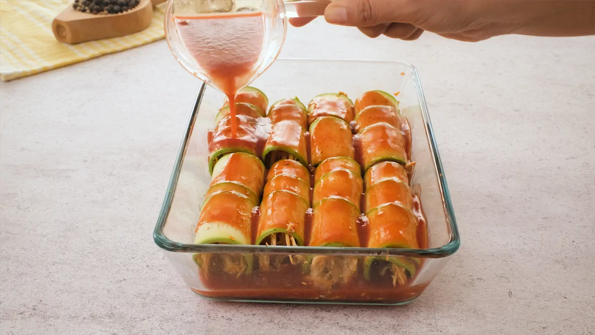 Zucchini rolls filled with chicken are arranged in a glass container, and tomato sauce is getting poured over them.