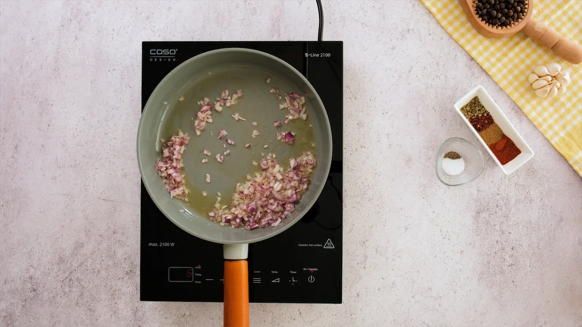 Red onions are getting sautéd in a pan, and the combination of spices required for this recipe is positioned next to the cooktop.