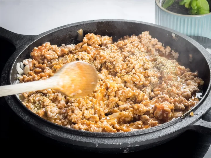 Mixing added spices into the meat sauce with a wooden spoon, cooking in a cast iron skillet.