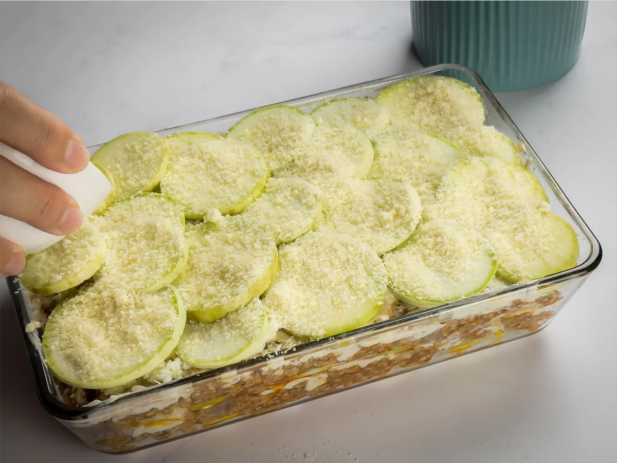 Sprinkling cheese over the top layer of zucchini slices before baking.