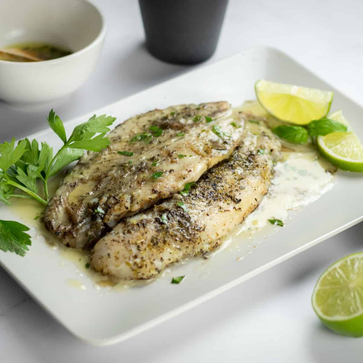 Pan-fried halibut filets with lemon butter sauce on a plate garnished with fresh green herbs and slices of lemon.