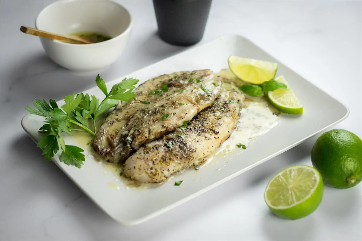 Pan-fried halibut filets with lemon butter sauce on a plate decorated with lemon and fresh green herbs.