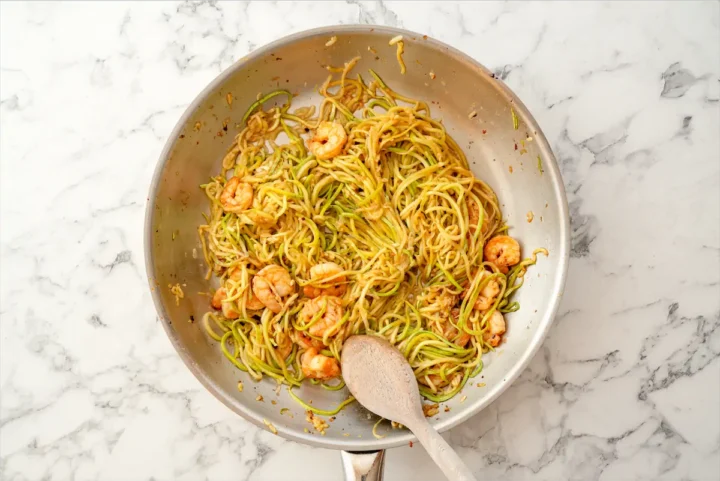 Zucchini noodles and shrimp is in a skillet with a wooden spoon.