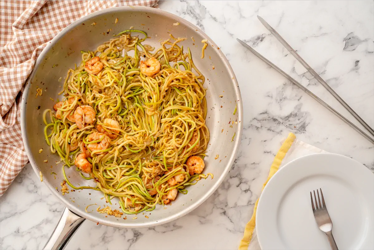 Shrimp zoodles in a stainless steel skillet on the table ready to be served with tongs, a fork and a plate .