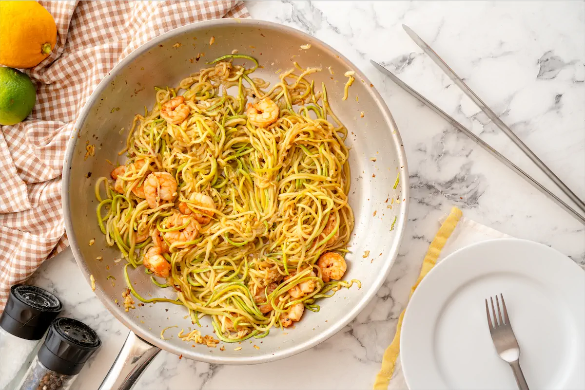 Tongs, a plate, and a fork are on the table to serve zoodles with shrimp from a steel skillet.