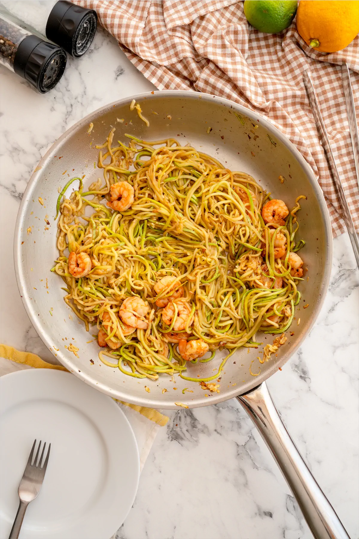 Zucchini noodles and shrimp in a steel skillet on the table beside a fork and a plate ready to be served.