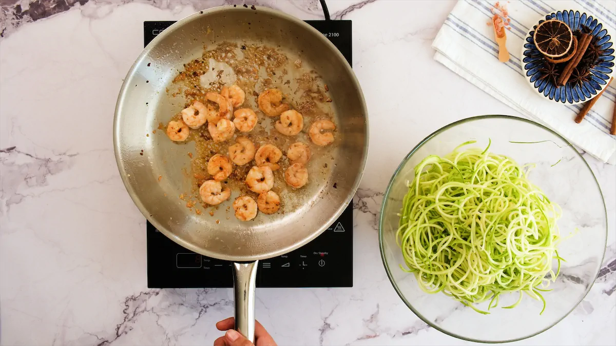 Sautéing shrimps in a stainless skillet on an induction stove beside a bowl of raw spiraled zoodles.