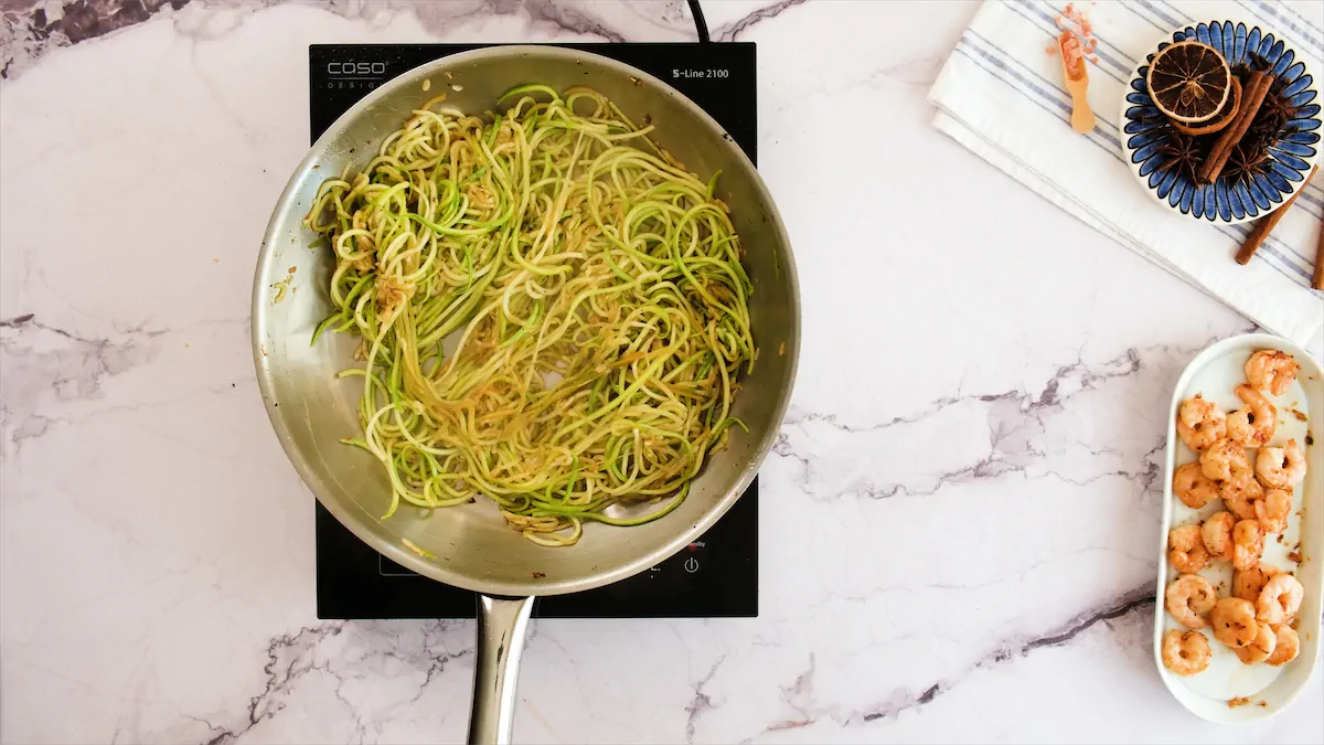 Cooking zucchini noodles in a stainless steel pan on an induction cooktop beside a plate of cooked shrimps.