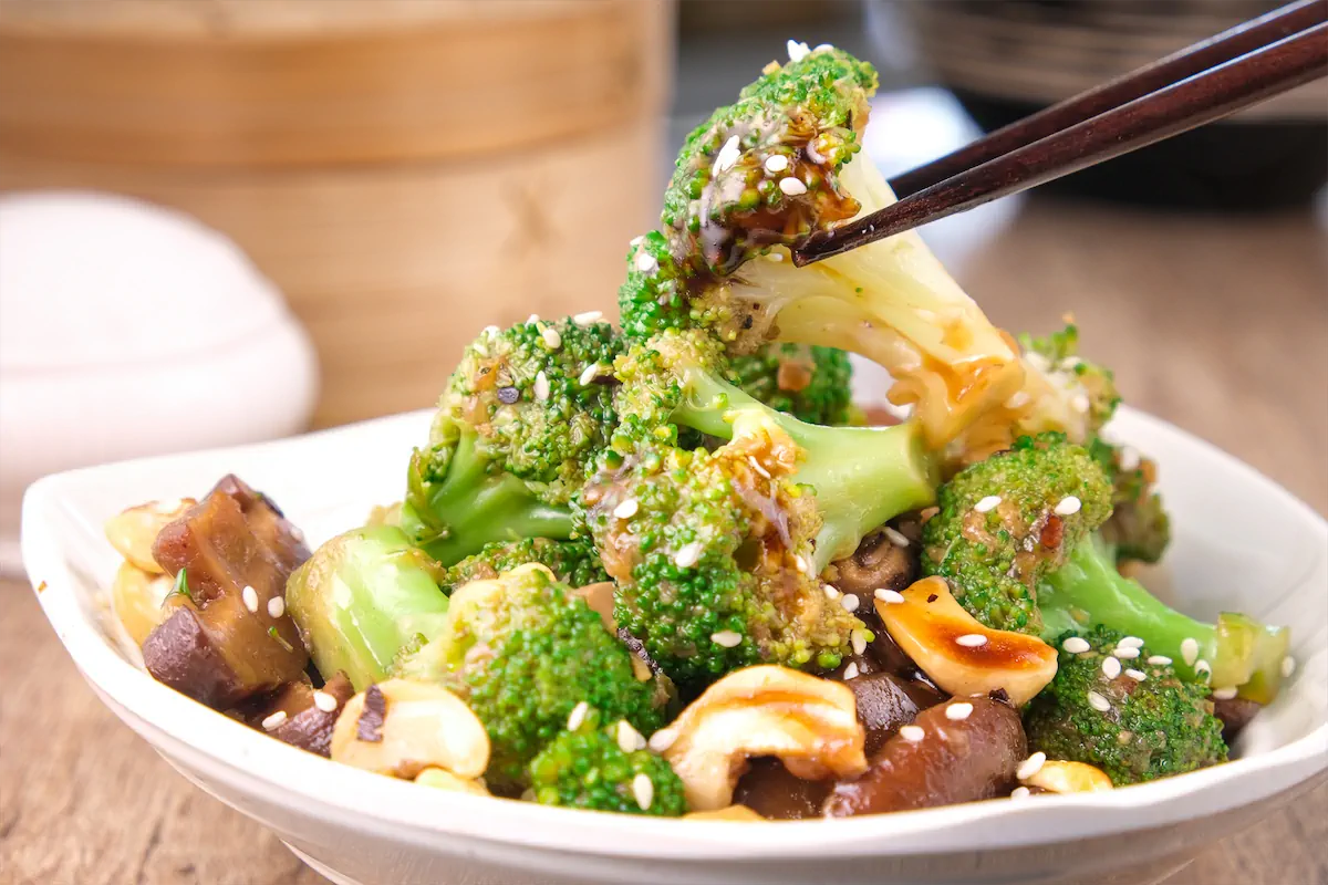 Keto broccoli and mushrooms stir-fried in an Asian sauce, with a piece of broccoli being picked up using chopsticks.