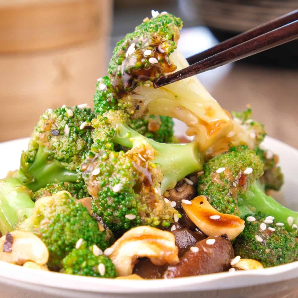 Stir-fried broccoli and mushrooms cooked in an Asian sauce, with a piece of broccoli being picked up using chopsticks.