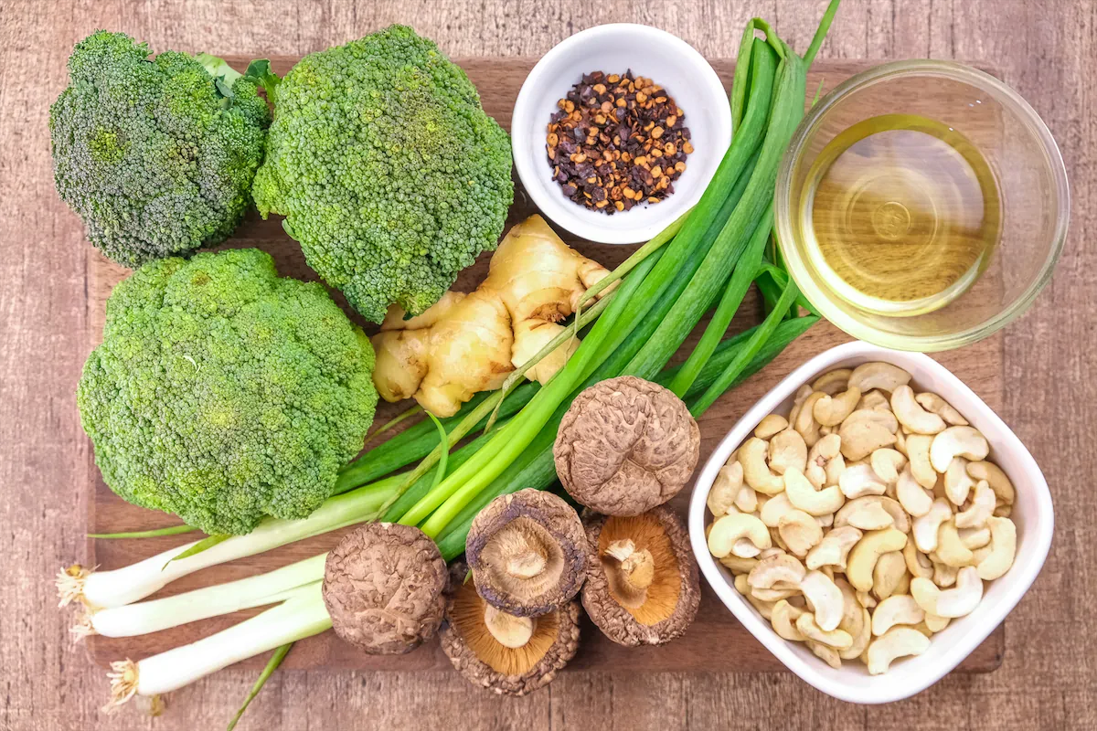 Dried Shiitake mushrooms, medium head of broccoli, canola oil, ginger, cashew nuts, and other required ingredients are arranged and displayed on the table.