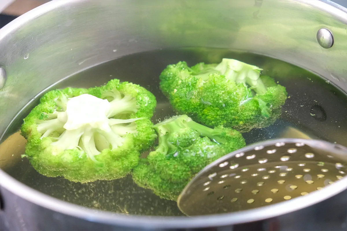 Broccoli is getting blanched in a pot of water using a steel perforated round spatula.