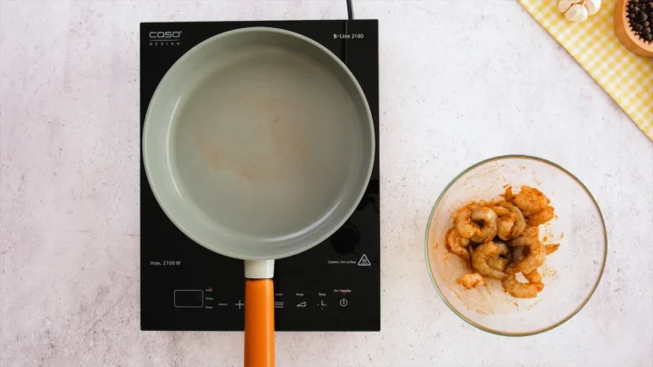A pan on an induction stovetop heating beside a bowl of marinated shrimp ready to be cooked.