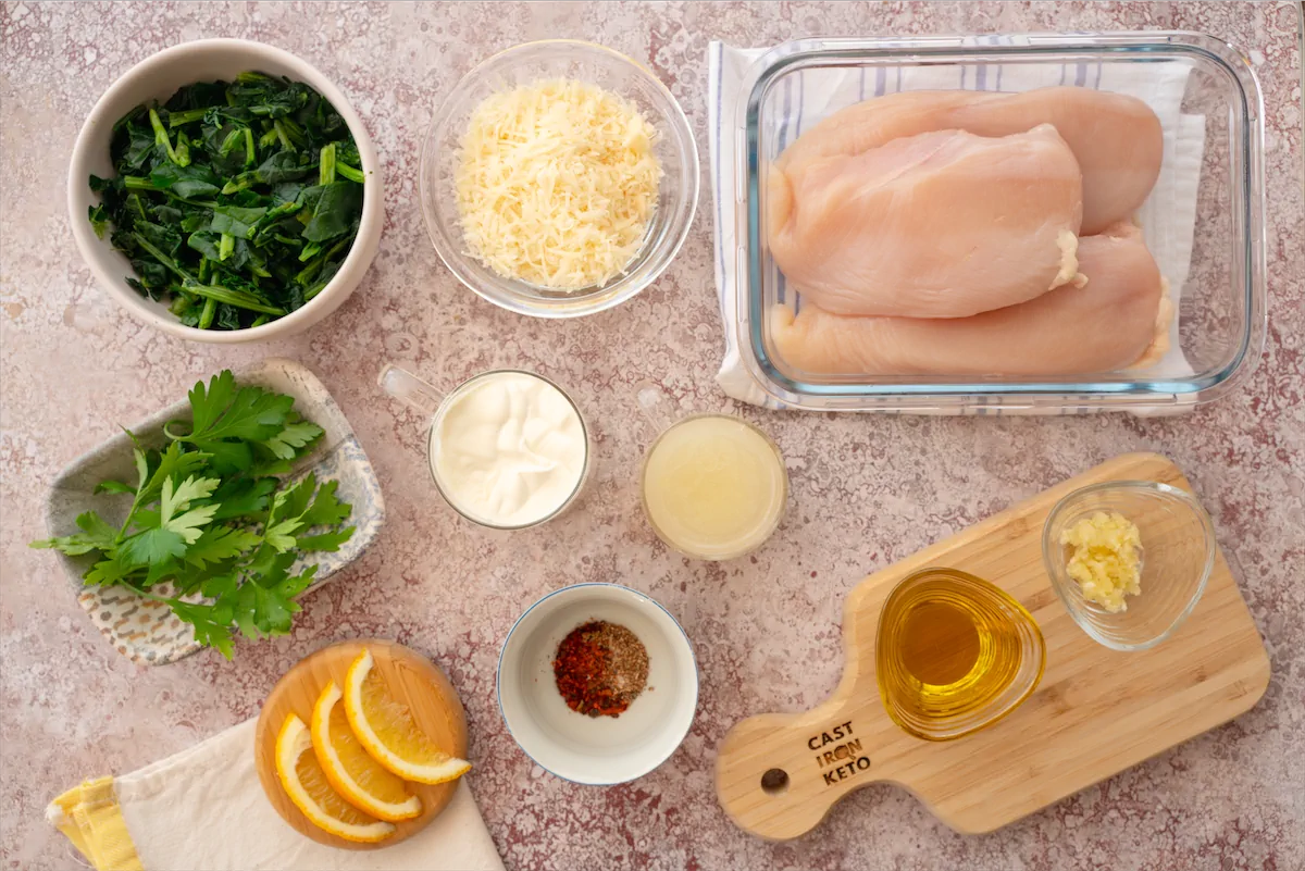 Skinless, boneless chicken breasts, avocado oil, garlic, minced, spinach, heavy cream, butter, grated Parmesan cheese, chicken broth, nutmeg, red pepper flakes, fresh lemon wedges for serving, fresh parsley, chopped for garnish arranged and displayed on the table.