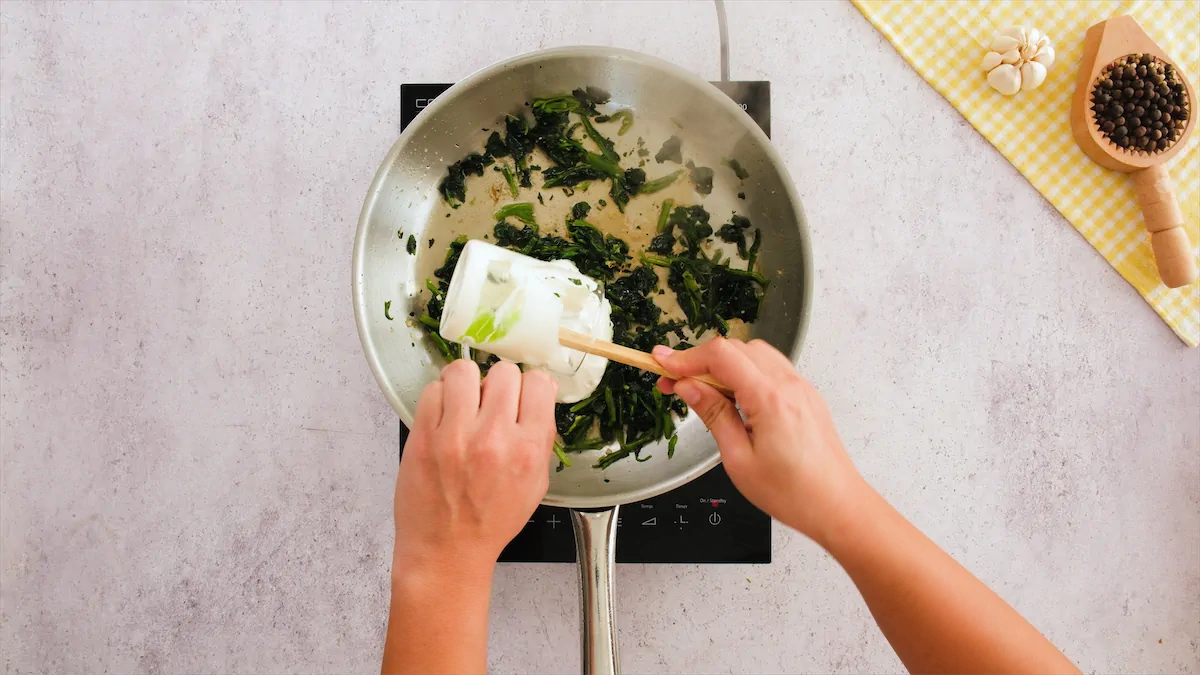 Pouring heavy cream to the skillet with spinach on an induction top.