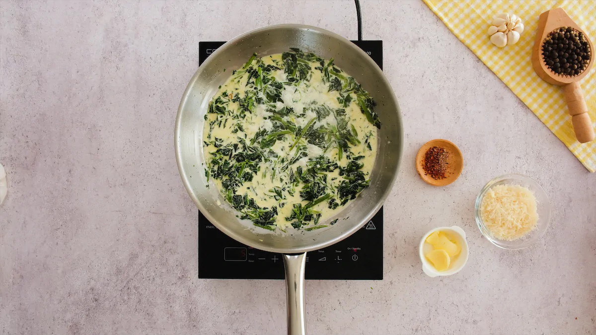 Letting the mixture of spinach, heavy cream and broth in a skillet to simmer beside Parmesan cheese, butter, nutmeg, and red pepper flakes in separate bowls.