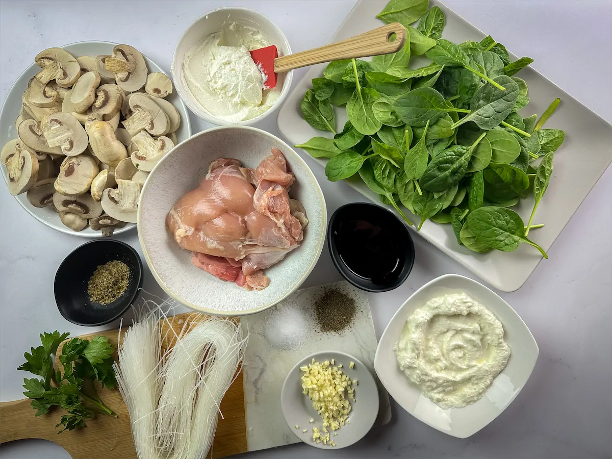 All the ingredients to make Keto chicken mushroom pasta including boneless, skinless chicken thighs, white button mushrooms sliced, konjac pasta, baby spinach, softened cream cheese, heavy cream, minced garlic, olive oil, dried parsley, freshly chopped parsley gathered and displayed on the table.