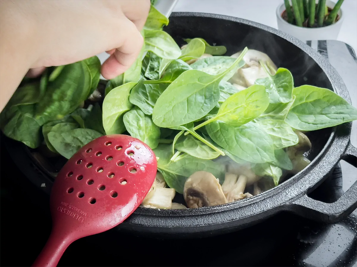 Fresh spinach added to the cast iron skillet with mushroom cooking on an induction stove top.