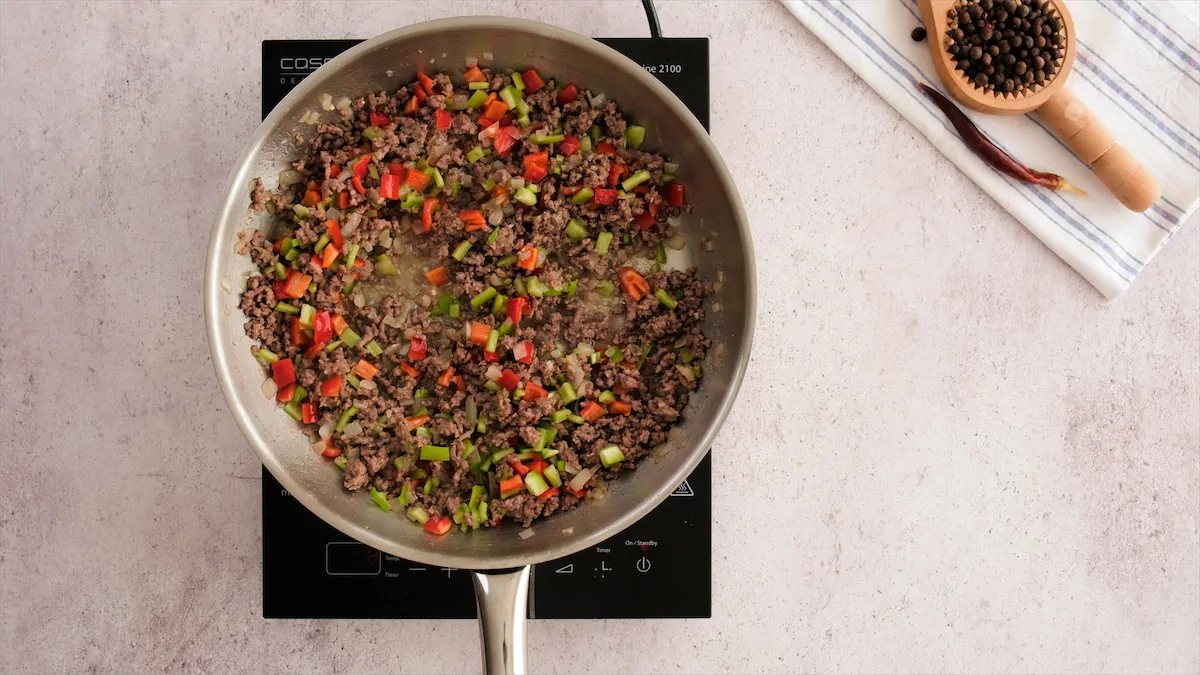 Ground beef cooking with chopped jalapeño and bell peppers in a stainless steel skillet.