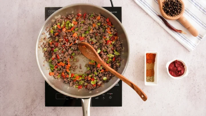 Stirring ground beef with a wooden spatula cooking with diced jalapeño and bell peppers in a stainless steel skillet.