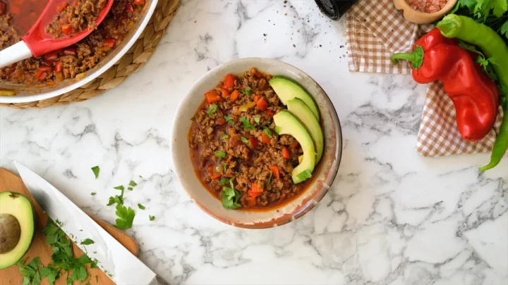 Homemade keto chili con carne in a bowl garnished with avocado slices and green herbs.