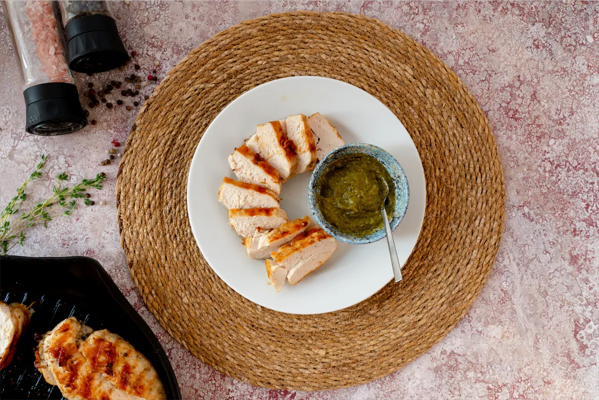 Slices of keto grilled chicken presented on a plate with a bowl of pesto sauce.