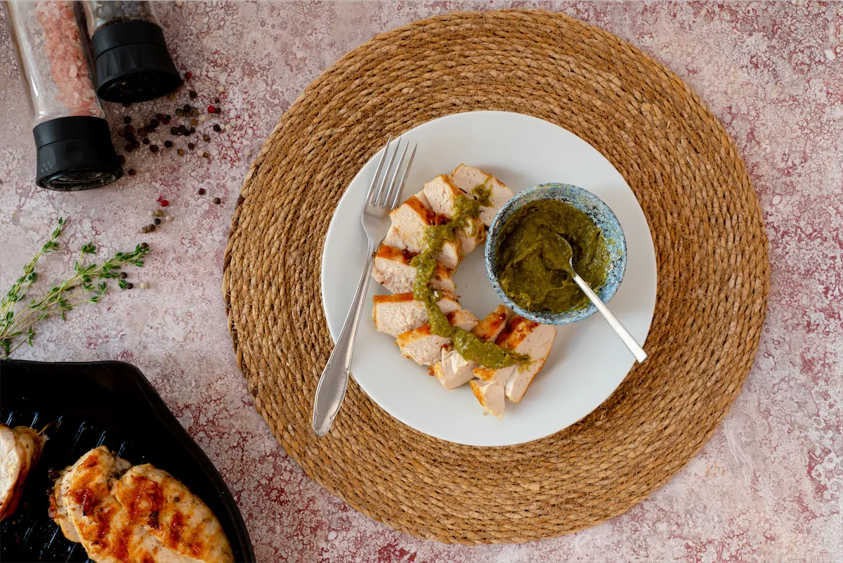 Slices of homemade grilled chicken on a plate, garnished with pesto sauce, and a side bowl of pesto sauce.