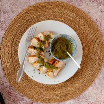 Slices of keto grilled chicken drizzled with pesto sauce served on a plate with a bowl of pesto sauce.