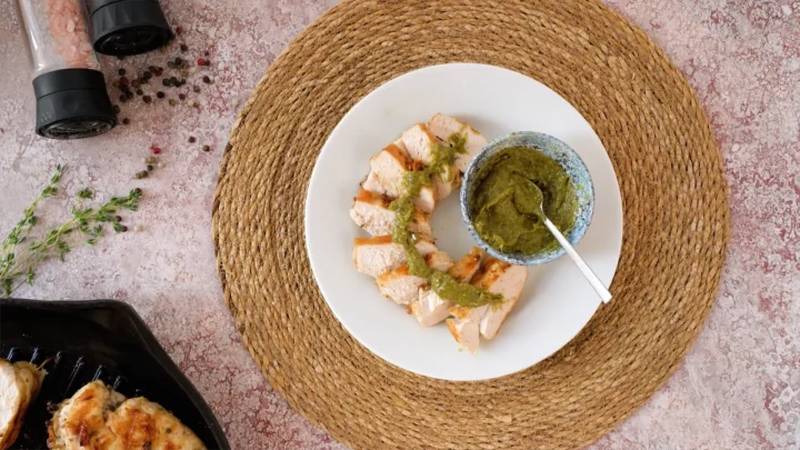 Keto grilled chicken slices topped with pesto sauce, arranged on a plate alongwith a bowl of pesto sauce.