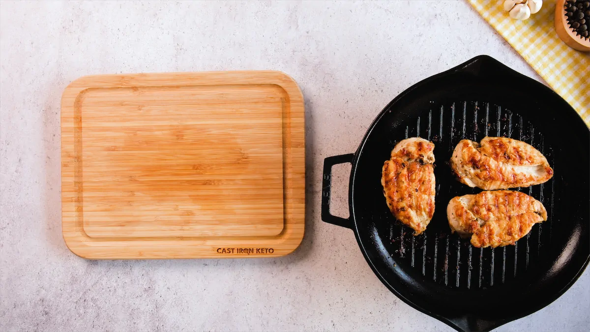 The grilled chicken filets are in a cast iron skillet beside a wooden board ready to be sliced and served.