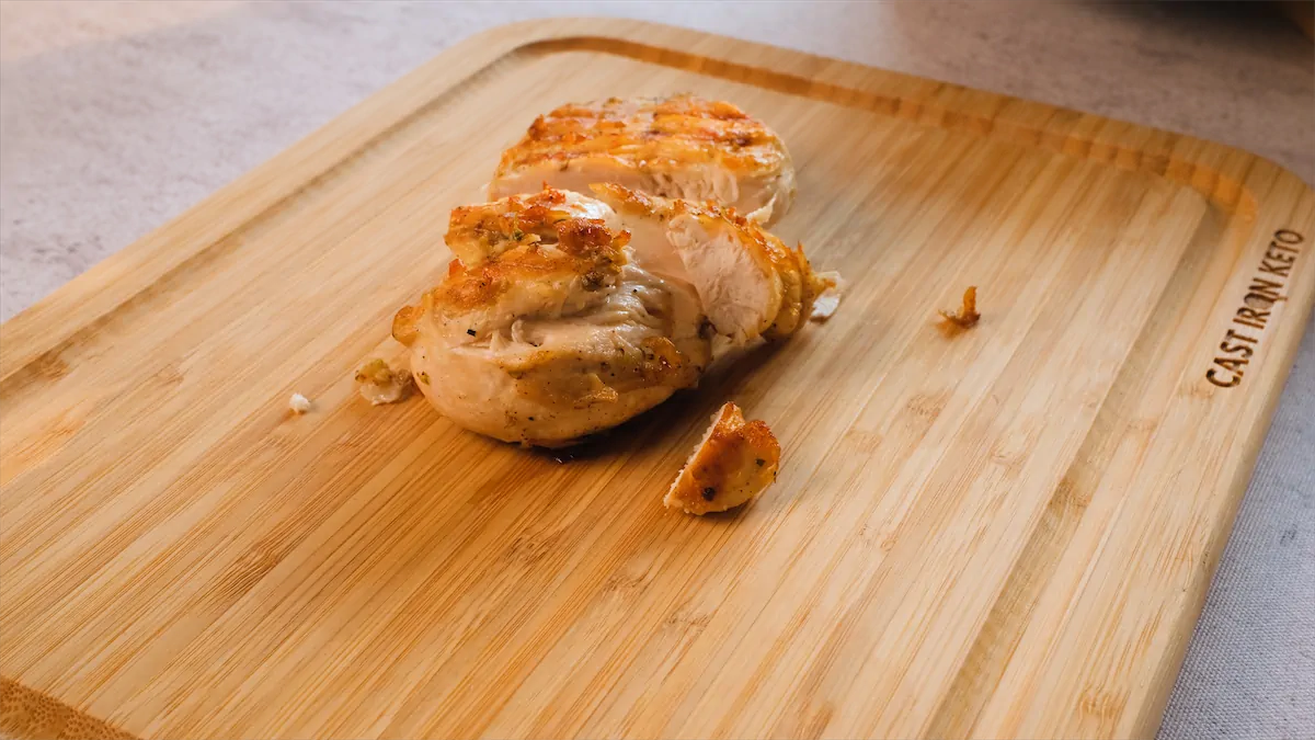 A filet of chicken is on a wooden board and it's getting sliced.