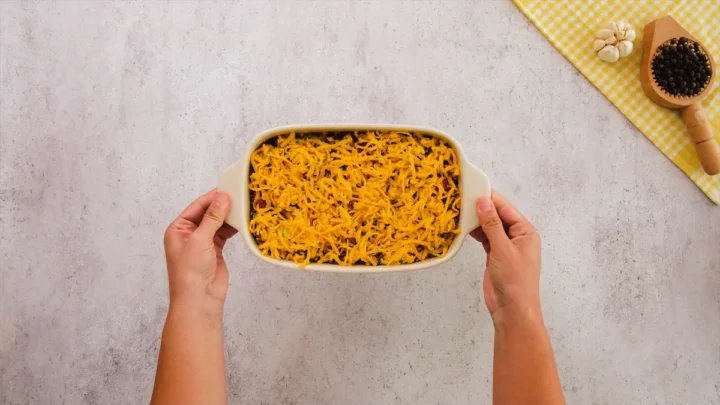 Two hands holding keto ground beef casserole in a baking dish topped with shredded cheddar cheese and ready to be placed in the oven for baking.