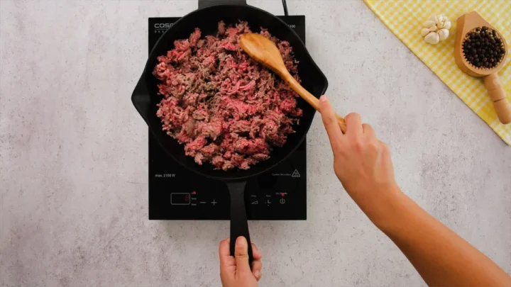 Browning ground beef in a cast iron skillet on an induction top and stirring it with a wooden spatula.