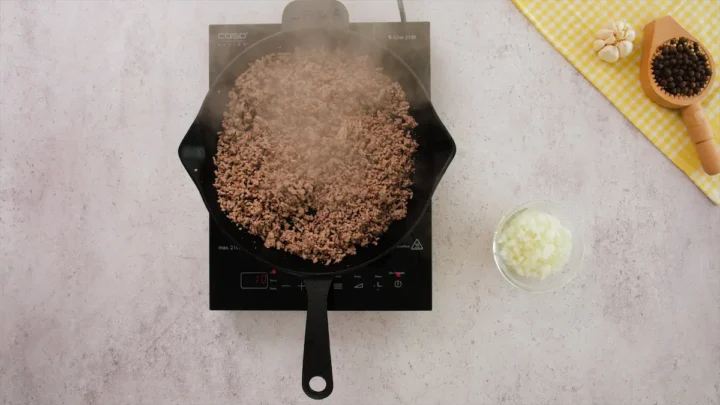 Ground beef is cooking in a cast iron skillet alongside a bowl of chopped onion.